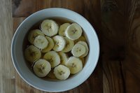 Fresh sliced bananas floating in clear liquid in white ceramic bowl on rustic wooden table, overhead view captured in natural lighting.