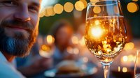 Bearded Caucasian man enjoying white wine at outdoor evening gathering with warm bokeh lights creating romantic atmosphere for dining and celebration.