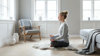 Woman in casual loungewear sitting cross-legged on fur rug in bright, minimalist bedroom, meditating or practicing mindfulness with serene expression, modern scandinavian interior design.