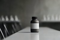 Black plastic bottle with blank white label on white table casting dramatic shadow, minimalist pharmaceutical or supplement packaging mockup in monochrome style.