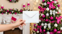 Hand holding white envelope against festive Christmas tree with pink and white ornaments, holiday greeting card delivery concept for seasonal celebrations.