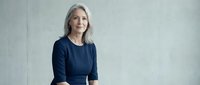 Professional mature Caucasian businesswoman with gray hair in navy blue dress sitting confidently against neutral background for corporate headshots.