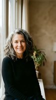 Confident mature woman with gray curly hair smiling by window in warm home interior for lifestyle and wellness content.