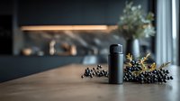 Sleek black thermos on wooden countertop with dark berries and greenery, showcasing modern kitchen design with blurred dark background.