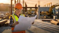 Construction worker in safety gear reviewing blueprints at building site during golden hour sunset with heavy machinery and structural framework.