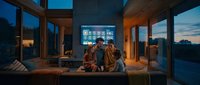 Young diverse couple relaxing on modern sofa watching streaming content on large TV screen in contemporary glass house during evening twilight hours.