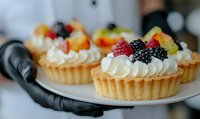 Pastry chef holding white plate with fresh fruit tarts topped with whipped cream and berries, ready for bakery, catering, or dessert menu use.