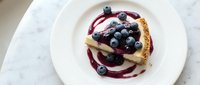 Gourmet cheesecake slice topped with fresh blueberries and berry sauce on white plate. Restaurant dessert presentation for menu photography and culinary content.