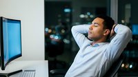 Young Asian businessman relaxing in office chair with hands behind head after work, city lights visible through window at night.