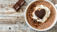 Hot chocolate topped with whipped cream and heart-shaped chocolate garnish in white ceramic mug on rustic wooden surface with chocolate pieces.