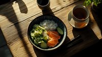 Fresh salmon sashimi bowl with cucumber, lettuce and rice served on rustic wooden table with green tea in morning sunlight, Japanese cuisine setting.