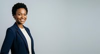 Young African American woman with short black hair wearing a dark navy blazer over a white shirt, smiling at camera against neutral gray background for corporate and business use.