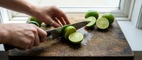 Chef hands slicing fresh limes on dark cutting board near window. Food preparation, cooking ingredient, culinary work, kitchen activity.