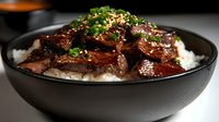 Glazed beef teriyaki slices served on white rice in black ceramic bowl, garnished with fresh chopped green onions and sesame seeds against dark background.