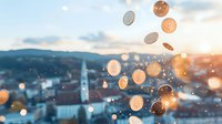 Falling golden coins against blurred cityscape with church spire at sunset, creating magical financial prosperity scene with bokeh light effects and sparkles.