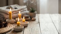 Lit candles in ceramic holders on wooden surface with spa accessories, towels, and plants in soft focus background for relaxation and wellness themes.