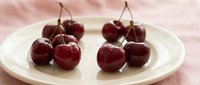 Fresh ripe red cherries with stems arranged on white ceramic plate against soft pink textile background for healthy eating and summer fruit concepts.