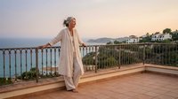 Senior woman in white flowing dress enjoying peaceful sunset moment on Mediterranean terrace overlooking azure sea and coastal resort town.