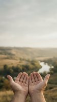 Open hands reaching toward scenic countryside landscape with river valley and rolling hills under cloudy sky for spiritual connection and gratitude concepts.