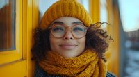 Young African American woman with curly hair wearing yellow knit beanie, glasses and matching scarf smiles warmly against yellow door background in soft autumn lighting.