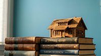 Miniature wooden house model sitting on stack of vintage books against blue wall background for real estate education concept.