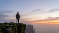 Man standing on rocky cliff edge overlooking misty valley at sunset, wearing backpack, contemplating scenic landscape during golden hour adventure travel.