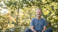 Middle-aged Caucasian man with beard sitting outdoors on moss-covered rock, wearing casual blue t-shirt, surrounded by blurred green foliage and natural sunlight, conveying relaxation and connection with nature.