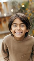 Young Latino boy wearing brown sweater smiling brightly outdoors with bokeh lights and autumn foliage in background, natural lifestyle portrait capturing genuine happiness.