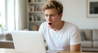 Young blonde man looking shocked and surprised while using laptop computer at home, expressing amazement at screen content in modern living room interior.