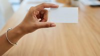 Woman with medium skin tone holding blank white business card in hand against wooden table background, suitable for corporate branding and professional networking materials.