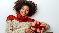 Young African American woman with curly hair wearing cozy knit turtleneck sweater holds wrapped Christmas present with red bow, expressing joy and warmth against white background.