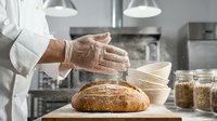 Baker in white chef coat dusting fresh artisan bread loaf with flour in professional kitchen bakery setting.