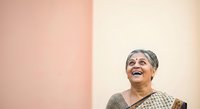 Senior woman with gray hair and warm smile wearing traditional patterned clothing against split pink and cream background. Suitable for lifestyle, diversity, and wellness content.
