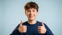 Happy teenage boy giving thumbs up gesture with both hands, smiling at camera against light blue background for positive youth content.