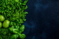 Fresh green herbs, limes, and leafy vegetables arranged on dark blue textured background with copy space, top view composition for food photography and menu design.