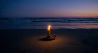 Vintage brass candlestick with burning candle on sandy beach at twilight. Ocean waves and purple sunset sky create romantic atmosphere for meditation.