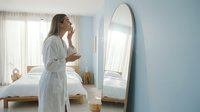 Young woman in white bathrobe examining face in oval mirror in bright bedroom with natural light, skincare routine and morning reflection concept.