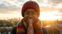 Young African American woman in colorful knit hat and scarf with hands in prayer pose smiling at golden hour sunset with city skyline background.