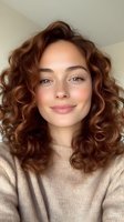 Young Caucasian woman with curly brown hair wearing beige sweater smiling naturally at camera with freckles and warm expression for lifestyle content.