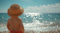 Young child wearing straw hat standing on beach looking at sparkling ocean waves under blue sky with white clouds during sunny summer day.
