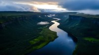 Dramatic aerial view of winding river through deep canyon valley with low clouds and sunbeams breaking through stormy sky, reflecting golden light on calm waters.