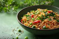 Asian noodles with vegetables and sesame seeds in dark ceramic bowl on green background, garnished with fresh herbs and chili peppers.