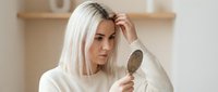 Young blonde Caucasian woman examining hair loss with hand mirror in modern bathroom, concerned expression, hair care and treatment concept.