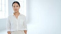 Young Asian woman wearing a light beige linen button-up shirt, standing in a bright minimalist interior space with natural window light, suitable for corporate and lifestyle content.