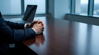 Professional businessman in dark suit with clasped hands at polished conference table in modern office boardroom with laptop and large windows.