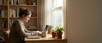 Man working on laptop at desk by window with plants and bookshelves, remote work and home office setup for business and productivity concepts.