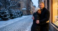Happy couple embracing on snowy winter street at dusk with warm lighting from shop windows creating romantic atmosphere for holiday season celebrations.