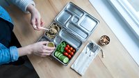 Caucasian woman in blue shirt preparing meal prep containers with fresh vegetables and grains on wooden surface for weekly lunch planning and healthy eating organization.