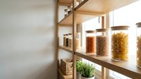 Wooden shelving unit displaying organized glass storage containers filled with dried pasta, grains, and legumes, with fresh herbs in a small pot, ideal for kitchen organization and pantry design projects.