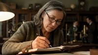 Mature woman with glasses writing in notebook at cozy cafe, focused on journaling or creative work in warm indoor setting with soft lighting.